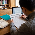 A journalism student, seen from behind, marks up a printed newspaper front page in red pen at a university library desk. The masthead reads The City Chronicle and the headline reads NEW RIVERSIDE PARK APPROVED. An open law textbook titled McNae's Essential Law for Journalists sits to the left, alongside a teal notebook and an open laptop, with bookshelves behind.