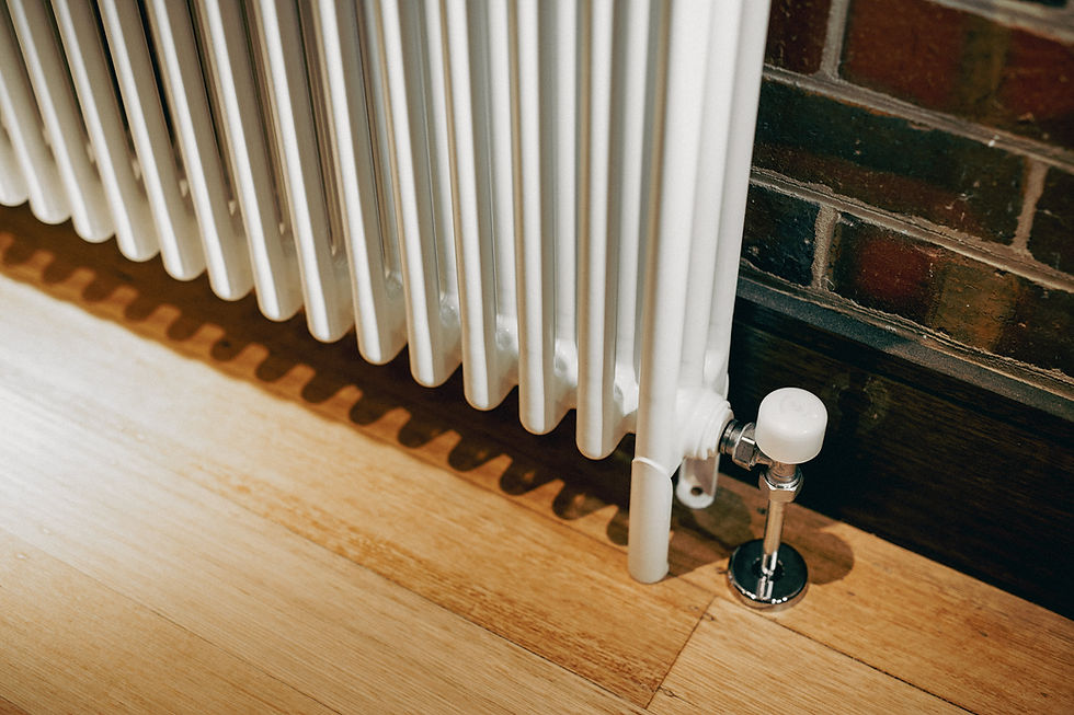 White radiator beside a brick wall on wooden floor. Chrome valve visible, casting shadows. Warm indoor setting.
