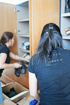 Two people organizing shoes into a wooden cabinet, one wearing a shirt with "Space & Order." Calm setting with cardboard boxes.