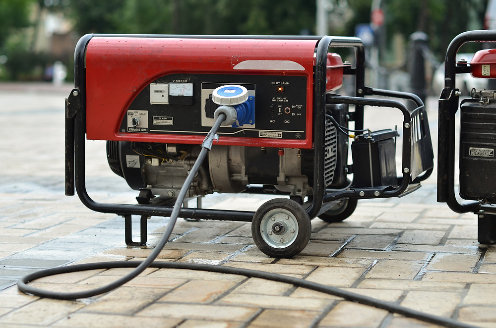 A generator with wires attached to it sits on a brick driveway at least 20 feet away from a home.