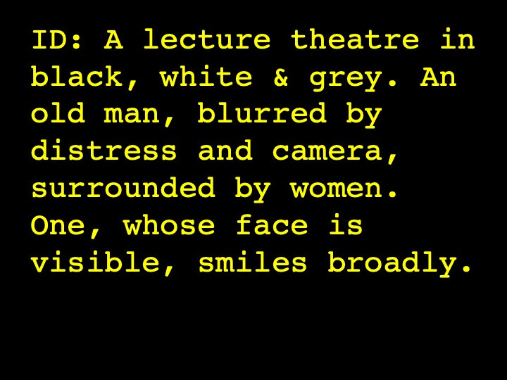 Image Description: A lecture theatre in black, white & grey. An old man, blurred by distress and camera, surrounded by women. One, whose face is visible, smiles broadly.