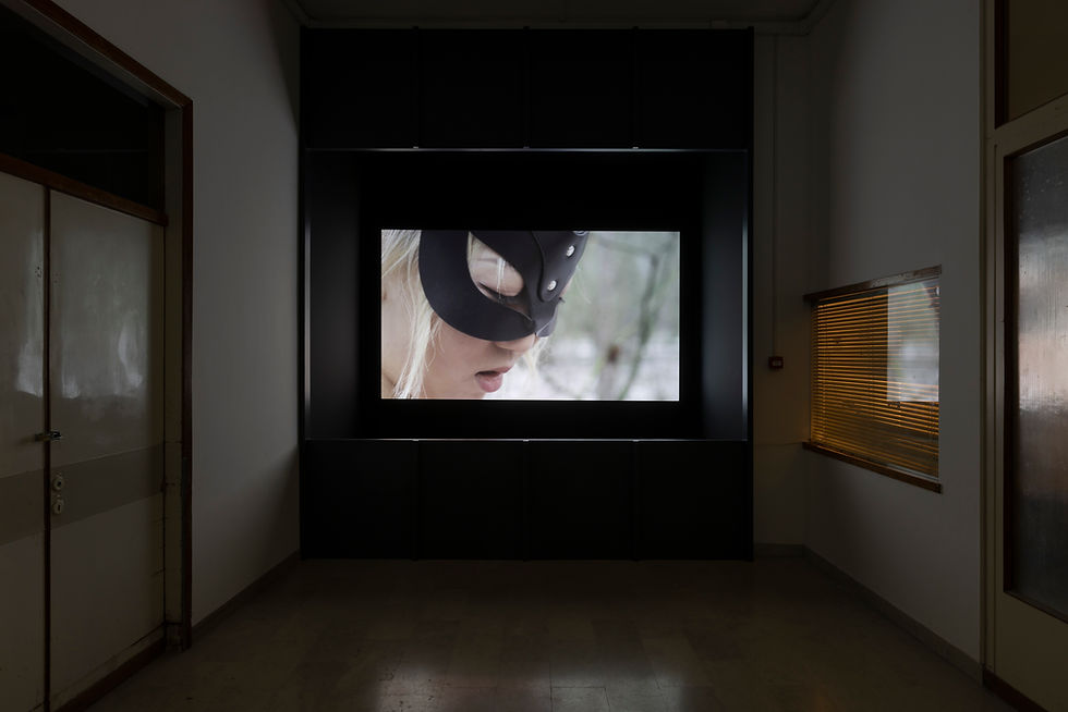 Photo of a dark installation room with a video depicting a young woman with bright blonde hair wearing a black leather mask and looking down.