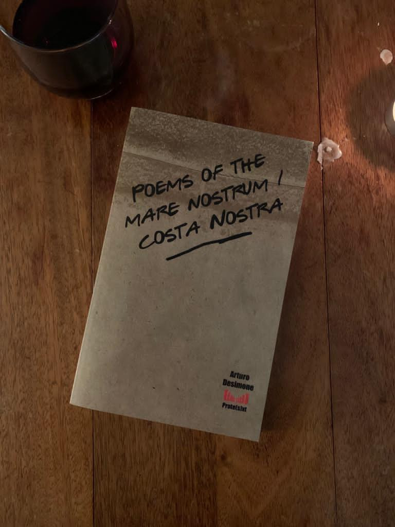 Photo of the pamphlet Poems of the Mare Nostrum/Costa Nostra, brown background with handwritten title in pen, on wooden surface with a glass of red wine in the left hand corner.