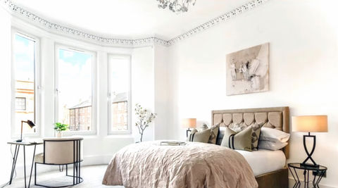 A luxuriously staged for sale master bedroom, with an office desk and chair, decorated in warm neutrals, creams with black accents, with an addedl office area in the bay window to highlight the space