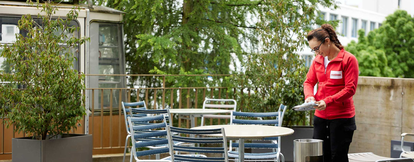 Das Bild zeigt eine Reinigungskraft der Prodoba Hotelservice AG, die mit einem Hochdruckreiniger die Terrassenmöbel eines Hotels reinigt. Sie entfernt Schmutz, Blätter und Verschmutzungen, um den Gästen einen angenehmen Aufenthaltsbereich im Freien zu bieten.