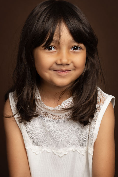 Close-up børneportræt – pige i hvid blondekjole smiler til kameraet, brun baggrund – studieportræt København