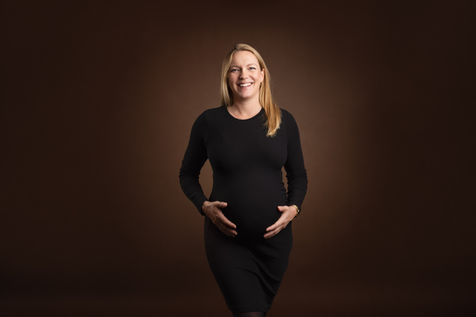 Gravid kvinde i sort, tætsiddende kjole står og holder om sin mave, smilende foran en brun studie­baggrund.