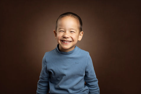 Glad dreng med et stort smil, iført en blå sweatshirt, siddende foran en varm brun studiebaggrund.