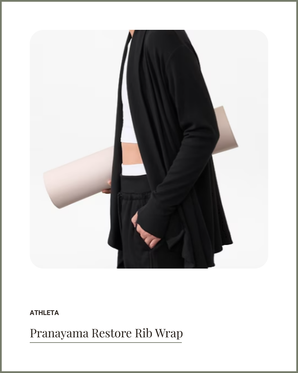 Person in a black wrap holds a yoga mat. White cropped top and black pants. Minimalist, calm vibe. Text: Athleta Pranayama Restore Rib Wrap.