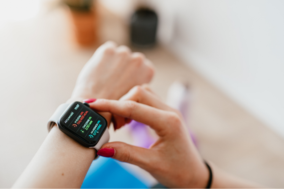 Close-up of a person adjusting a smartwatch displaying fitness stats. Bright nails, blurred indoor setting with soft colors.
