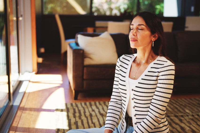 Woman meditating on patterned rug in sunlit room, wearing striped cardigan. Brown sofa with cushions in background, calm atmosphere.