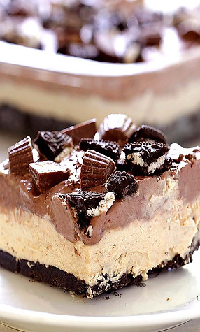Close-up of a creamy peanut butter dessert bar topped with chocolate, Oreos, and peanut butter cups on a white plate.