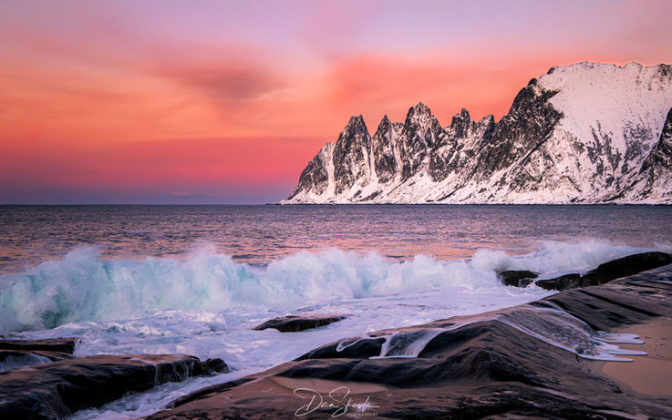 Vakre bølger bryter mot steinene ved kysten med fjell i bakgrunnen.