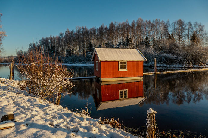Red house by the water with snow and trees in the background. Atelie Melheim.