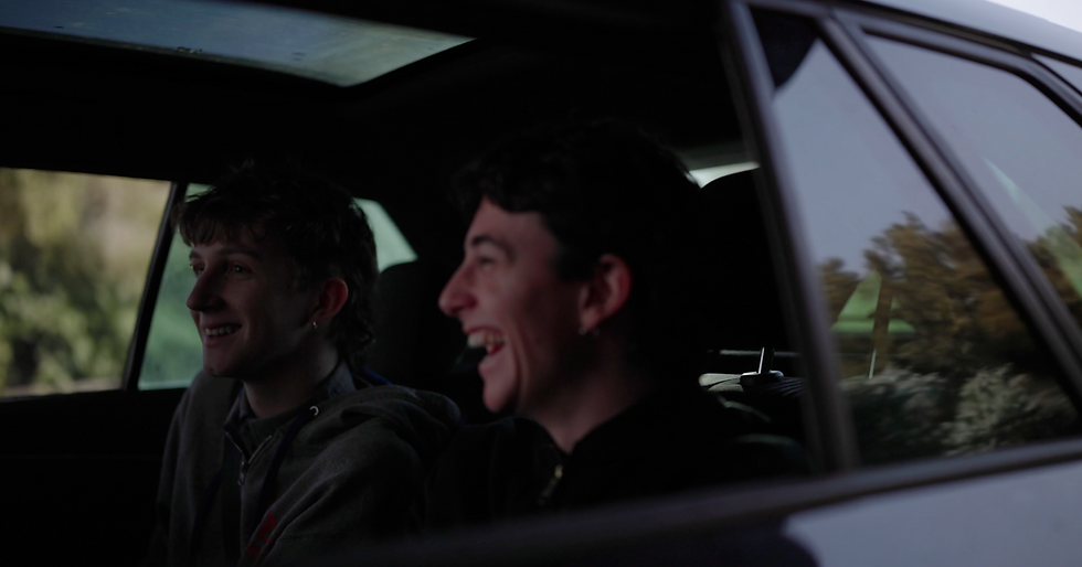 Two people are sitting in a car, smiling and laughing. Sunlit trees are visible through the windows, creating a relaxed, joyful mood.