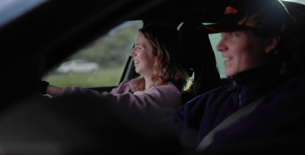 Two people smiling inside a car, one driving and the other in a cap. Bright, grassy background, mood is happy and relaxed.