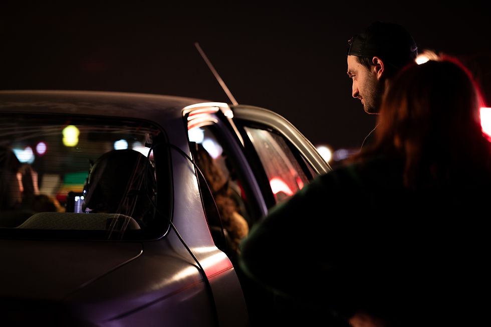 Man in cap leans into car with people inside at night, illuminated by red and yellow lights. Dark, urban setting with a focused mood.