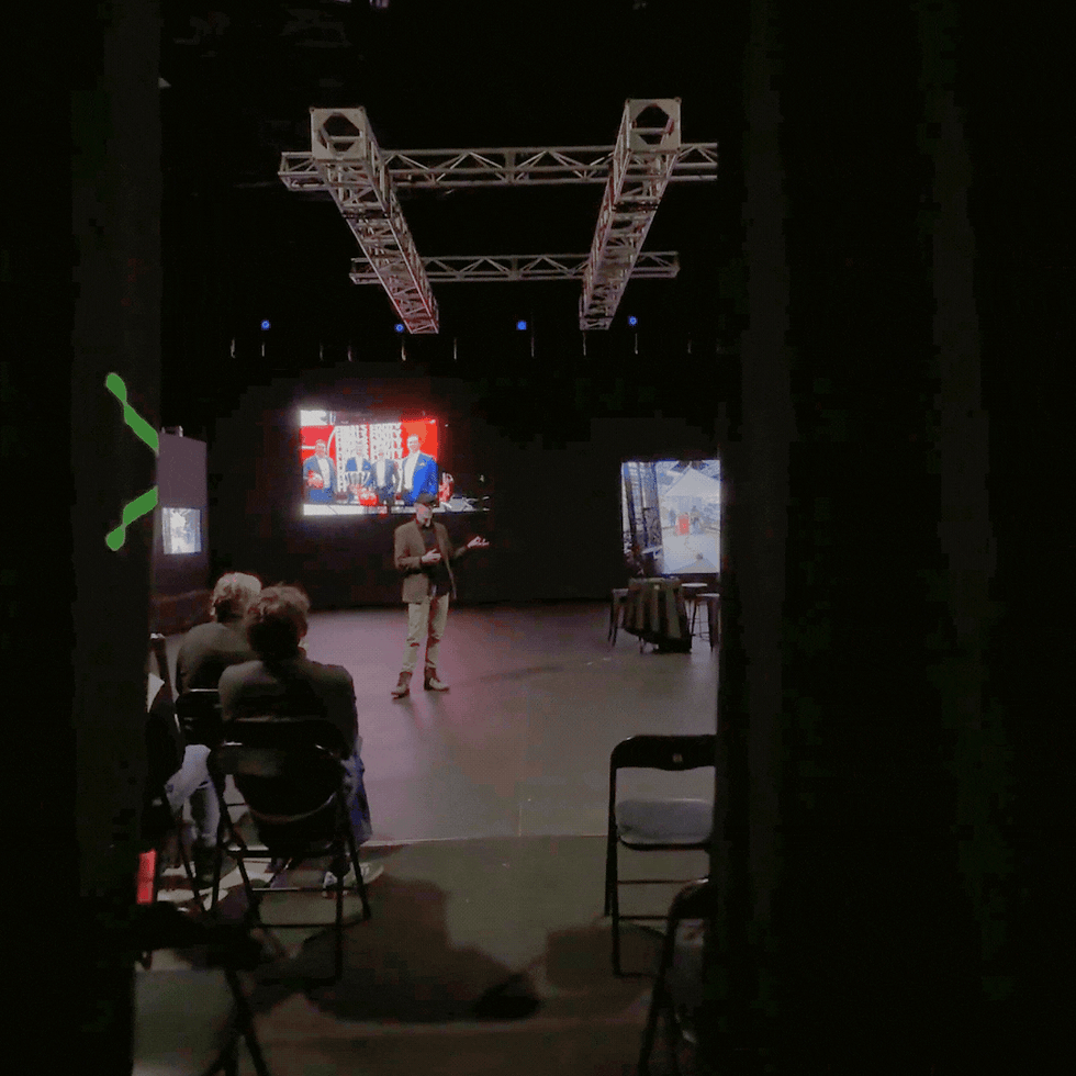 A person onstage presents to a seated audience in a dimly-lit theater. Screens display colorful images, creating an engaging atmosphere.