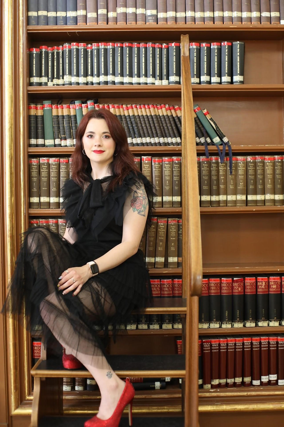 Woman in black dress and red heels sits on library steps, surrounded by books. She smiles confidently, with a tattoo on her arm.