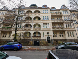 A cream Berlin apartment building in Steglitz with rows of arched balconies and bay windows behind bare autumn trees, cars parked along the street in front
