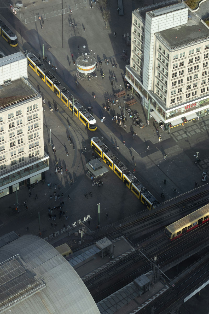Aerial view of a city square with yellow trams, people walking, and modern buildings. A train is visible on tracks below. Shadows cast across the scene.