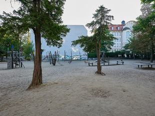 A quiet morning at the playground on Rheingaustrasse in Berlin-Friedenau, sand and climbing frames under mature trees with a pale apartment gable behind