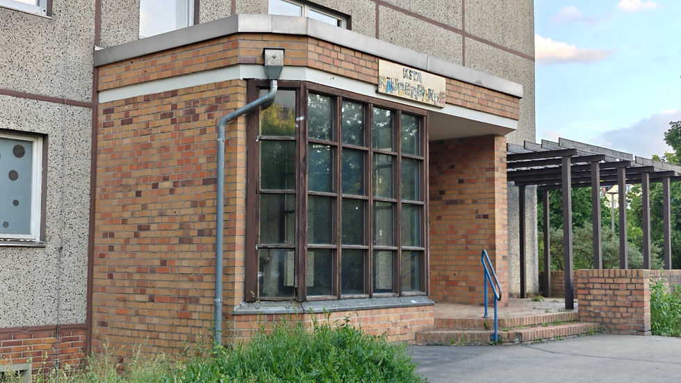A closed-up brick corner entrance of a former Kita in a housing block at Ernst-Thaelmann-Park in Berlin, faded lettering above a large timber-framed window, an empty pergola walkway to the right and grass growing untended at the base