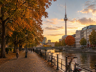 Riverbank lined with autumn trees, people walking and cycling. TV tower and buildings in background under a colorful sunset sky. Calm mood.