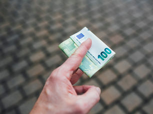 Hand holding a folded 100 euro note with cobblestone pavement background. The note is green and white, featuring the number 100.