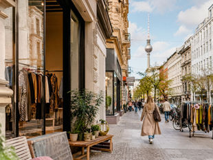 Woman in beige coat walks down a Berlin shopping street past clothing racks, with the TV Tower in the background.