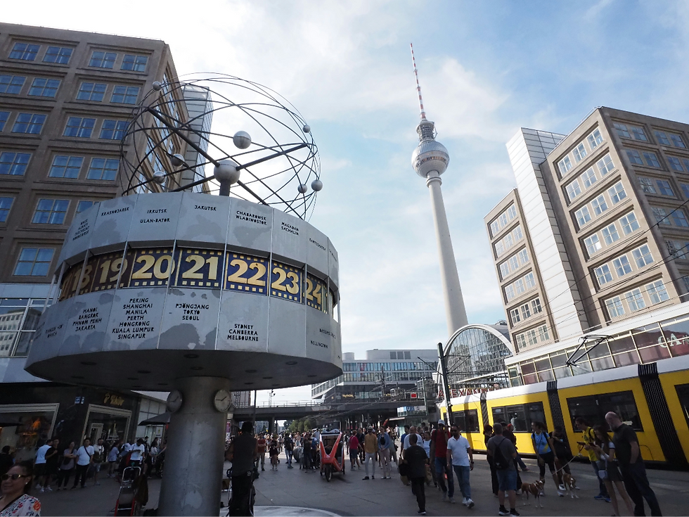 People walk by the World Clock and yellow tram in Alexanderplatz, Berlin, with the TV tower in the background under a blue sky.