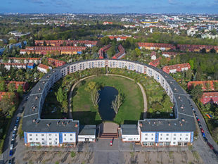 An aerial view of the Hufeisensiedlung in Berlin-Britz, the horseshoe-shaped white housing block curving around a green courtyard and a small pond, with rows of red-roofed terraced houses beyond