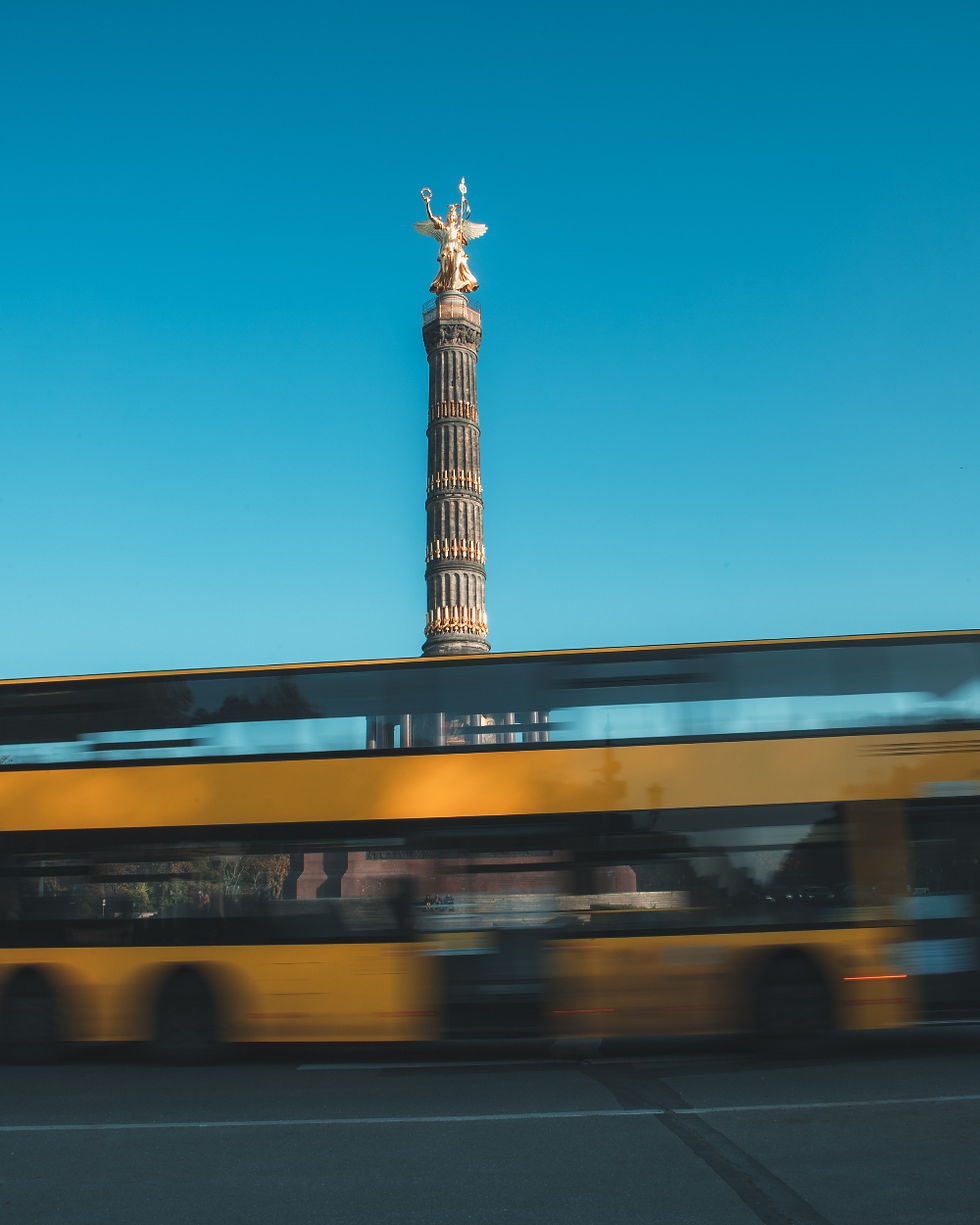 A yellow double-decker bus passes in front of the Victory Column statue against a clear blue sky, creating a dynamic urban scene.