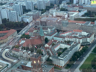 Aerial view of a dense European city with a red-roofed church, clock tower, and winding roads beside tall apartments.