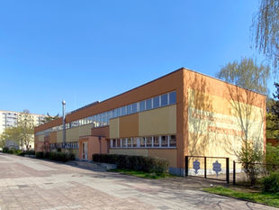 A long two-storey Kita on Otto-Marquardt-Strasse in Berlin-Lichtenberg under a clear blue sky, orange and yellow render, the words Kindertagesstaette Entdeckerland painted large on the end wall, Plattenbau housing behind