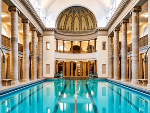 Ornate indoor pool with blue water, surrounded by tall stone columns and a domed ceiling with mosaic patterns. Lifebuoys are visible.