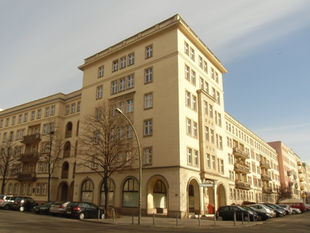 A corner block on Weidenweg in Berlin-Friedrichshain under a clear blue sky, five storeys of pale render above an arcade of ground-floor openings, bare trees along the pavement and cars parked in front