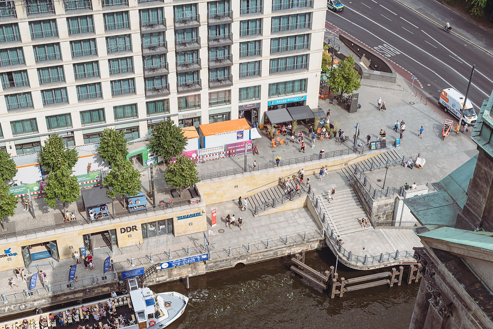 Aerial view of the DDR Museum on the Spree riverbank in central Berlin, with tour boats passing below and the Berliner Dom in the foreground