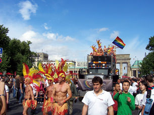 Feathered dancers and a rainbow-flag truck at a crowded pride parade near the Brandenburg Gate under a bright blue sky