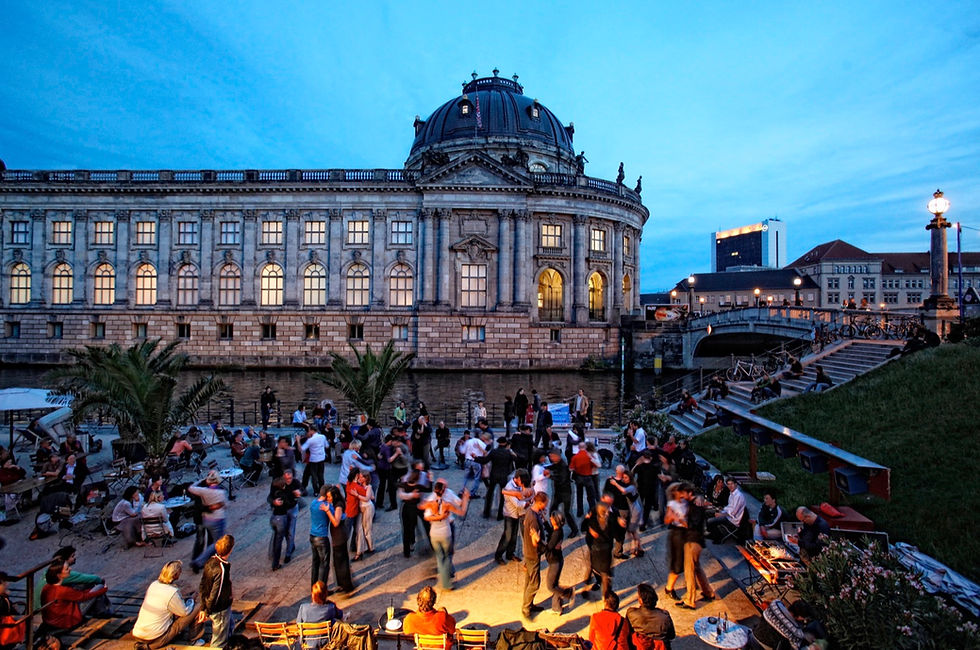 People at riverside tables at Strandbar Mitte on the Spree in Berlin on a summer evening, with the Bode Museum lit up behind