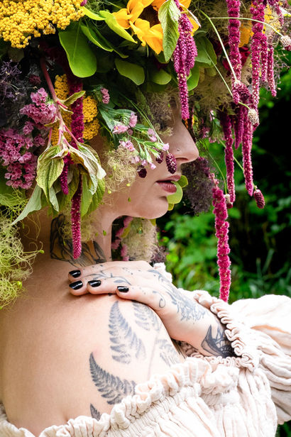 Künstlerisches Farbporträt einer tätowierten Frau im Profil mit einem opulenten floralen Kopfschmuck aus gelben und pinkfarbenen Blüten, hängenden Blütenrispen und üppigem Grün. Sie trägt ein cremefarbenes Kleid, legt eine Hand auf ihre Schulter und steht vor einem grünen, unscharfen Naturhintergrund.