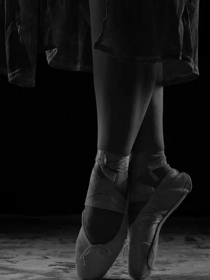 Schwarzweiss Detailaufnahme von Unterschenkel und Füße in Pointe-Schuhen. Die Bänder sind um die Knöchel gebunden. Dunkler Hintergrund, feine helle Partikel am Boden sichtbar.