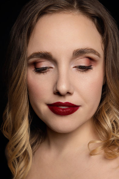 Professionelles Beauty-Porträt einer Frau mit geschlossenen Augen, schulterlangen, braunblonden Haaren und sanften Locken. Ihr Make-up kombiniert schimmernden rosafarbenen Lidschatten, einen feinen Lidstrich, lange Wimpern und kräftige dunkelrote Lippen. Das Gesicht ist vor einem tiefschwarzen Hintergrund weich ausgeleuchtet.
