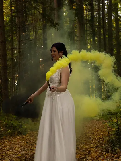 Farbaufnahme im Wald. Frau im Brautkleid in seitlicher Position. Gelber Rauch zieht in einem Bogen hinter ihr her. Eine Rauchfackel ist in der Hand sichtbar. Wald und Herbstlaub im Hintergrund.