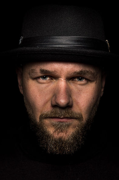 Künstlerisches Low-Key-Porträt eines bärtigen Mannes mit schwarzem Hut und intensivem, direktem Blick. Warmes Licht hebt sein Gesicht und seine hellen Augen vor tiefschwarzem Hintergrund hervor.