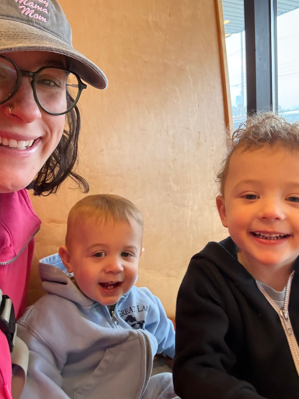 Three people smiling indoors: a person in a pink hoodie and cap, a toddler in a light blue hoodie, and a child in a black hoodie. Cozy mood.