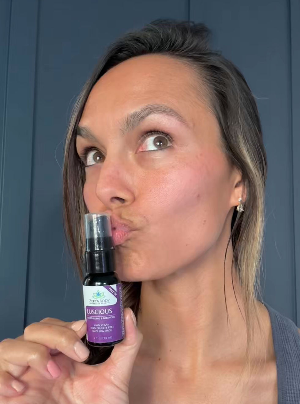 Woman kissing a small bottle labeled "Luscious Face Oil" with a purple and white design. Blue wall background, playful mood.
