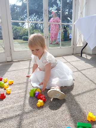 A toddler in a white dress plays with colorful toy blocks on a sunny floor inside a room with glass doors