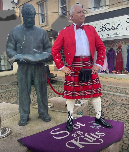 Man dressed in a vibrant red formal jacket and red tartan kilt standing next to a stone statue outdoors
