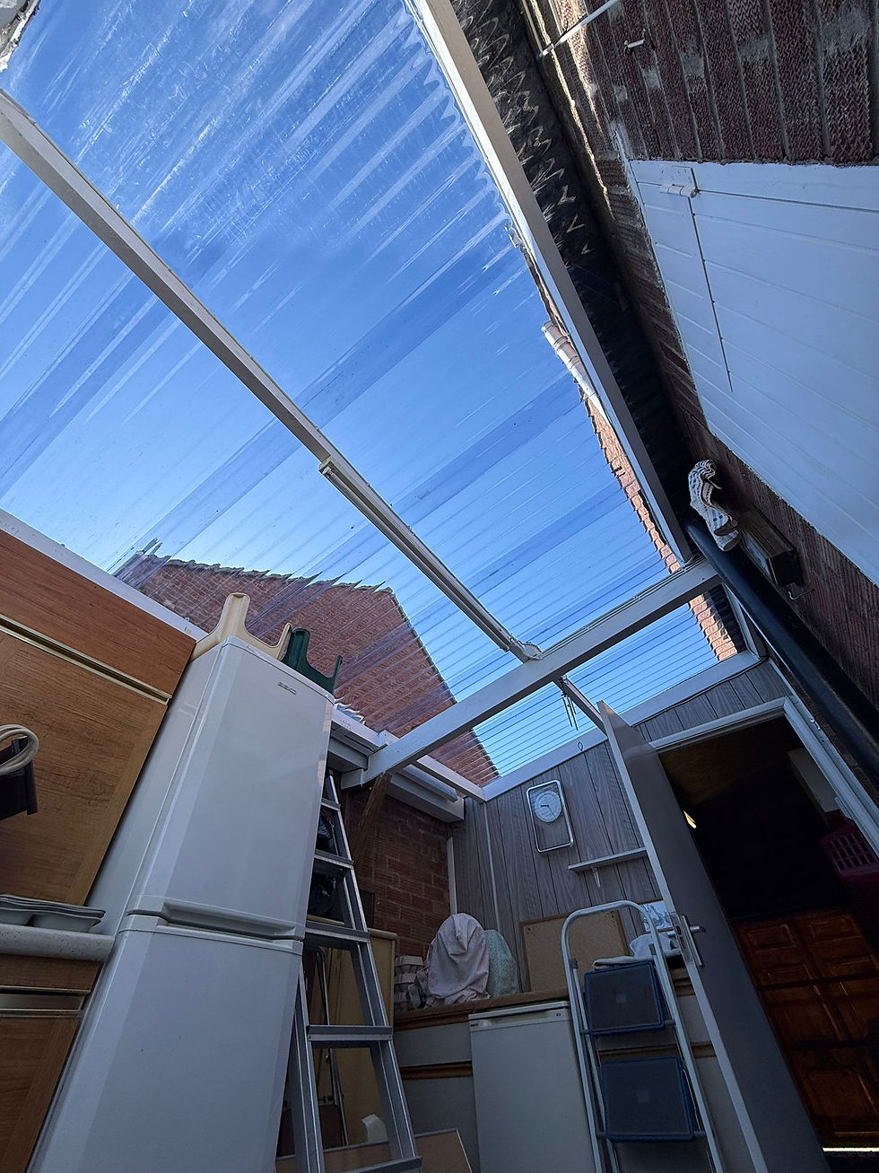A low-angle view of a lean-to side extension with a clear corrugated roof and storage items
